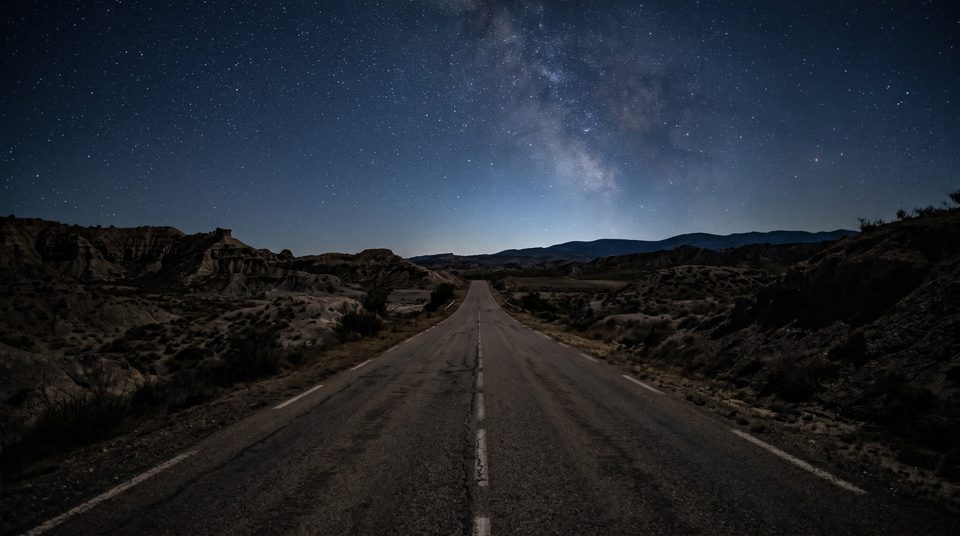 Route droite qui traverse un paysage aride sous un ciel étoilé, avec une ligne d'horizon bleutée